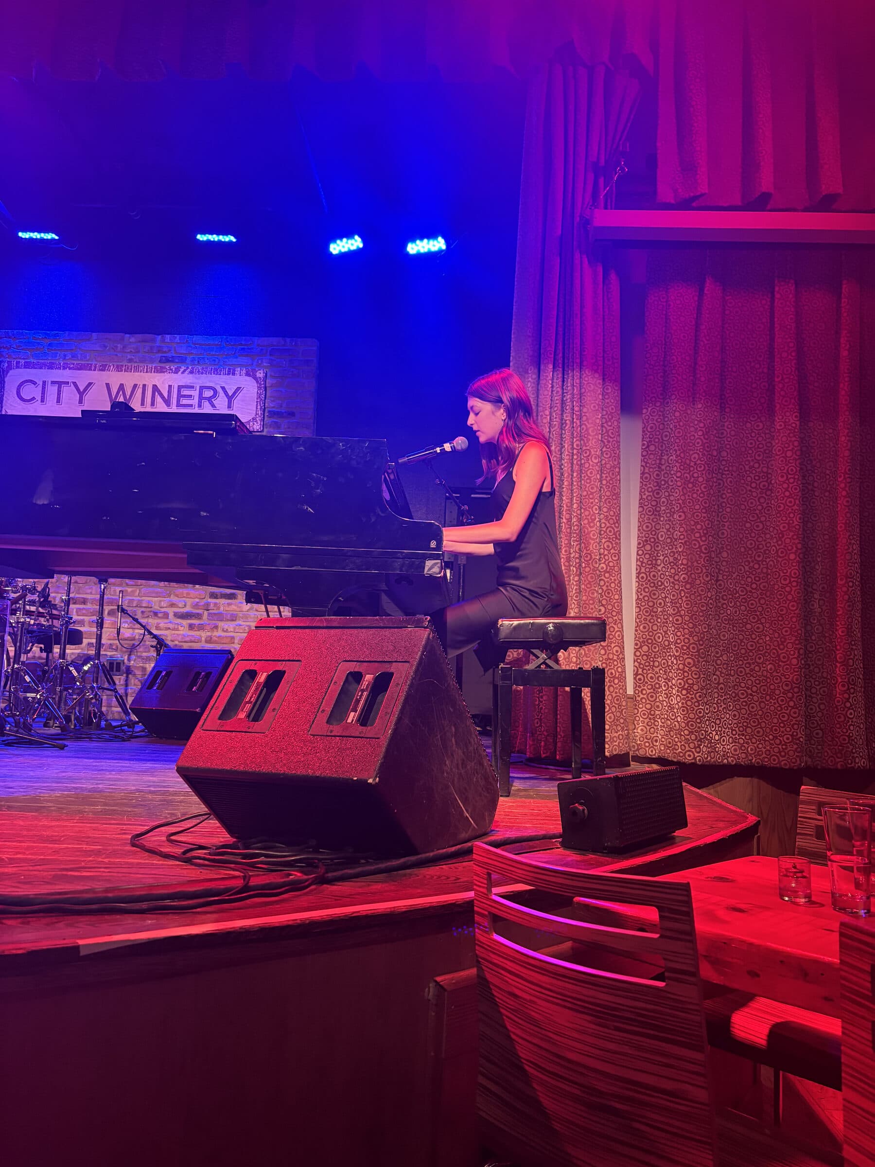 Thelma sings into a microphone at a grand piano, lit red and blue, on the City Winery stage in Chicago.